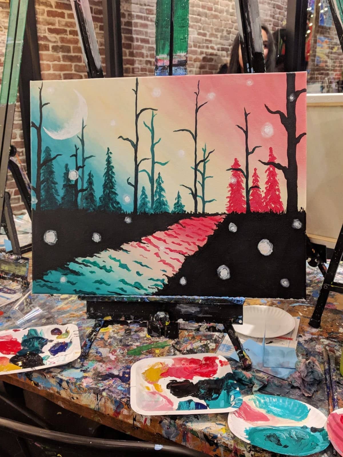 Painting of a gradient sky from teal to coral with silhouettes of trees and the sky reflected in the river. Snowflakes pepper the landscape, and there's a large crescent moon. Painted at Muse Paintbar